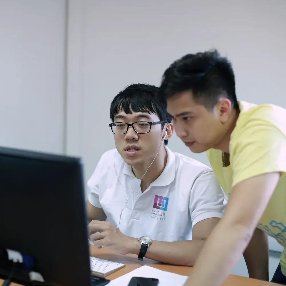 Two Eastgate engineers pairing at one monitor, one leaning in to point at the code
